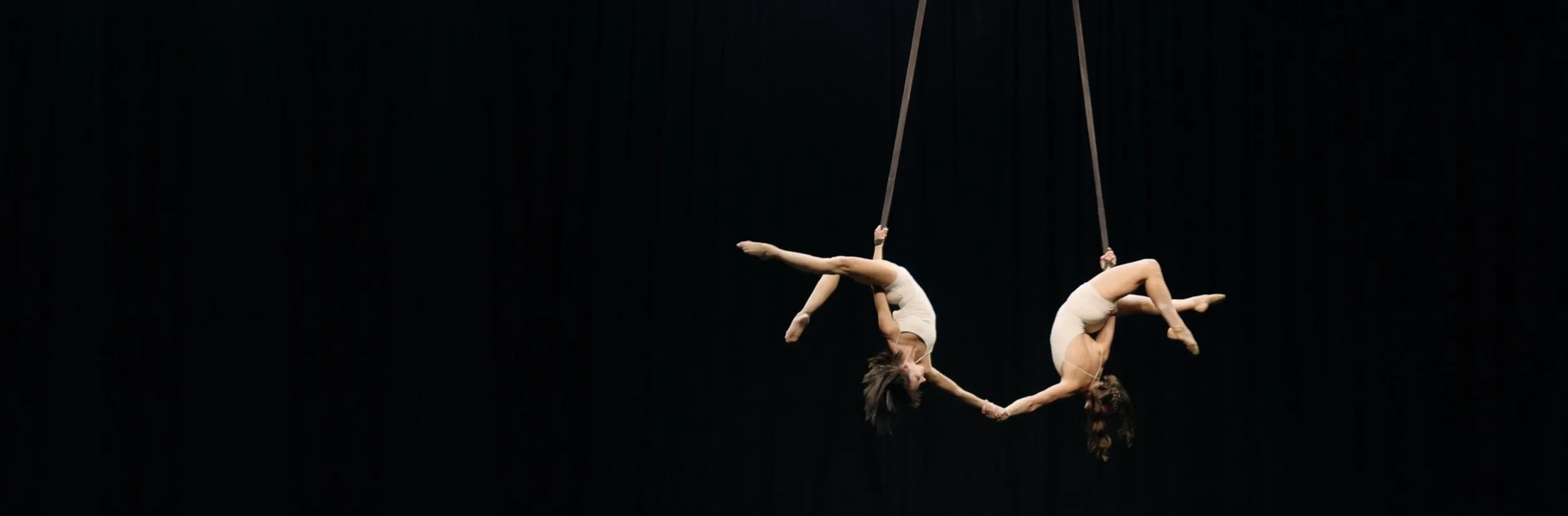 Charlee Shae and Sara Knauer suspended on aerial straps, hand in hand, on a darkened stage.